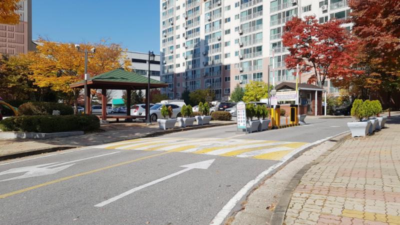 차량 출입차단기가 가운데 설치되어 있어 외부 차량의 출입을 통제하고 있으며, 경비실은 안쪽에 위치해 있어요